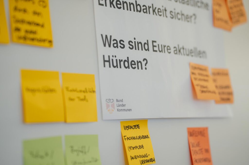 A sheet at a wall with 2 questions; the question in focus asks: “What are your current barriers?” with a black-red-golden government eagle symbol of federal, state and local government; various sticky notes are put up around the sheet, one reads: “out-dated case-working systems and complex connection procedures”