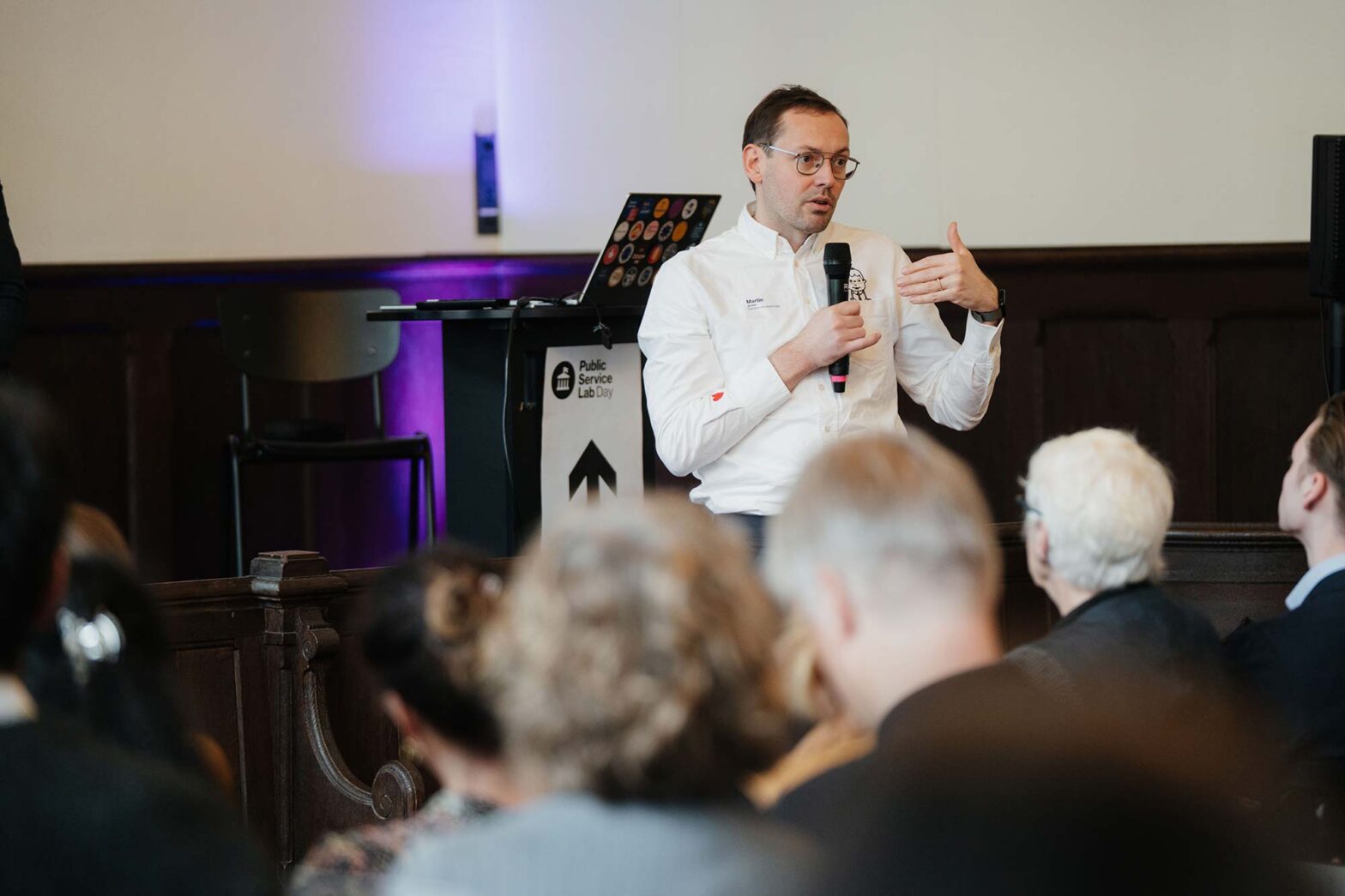 A light-skinned middle-aged man wearing a white shirt holding a microphone, standing in front of a lectern, speaking in fron of an audience