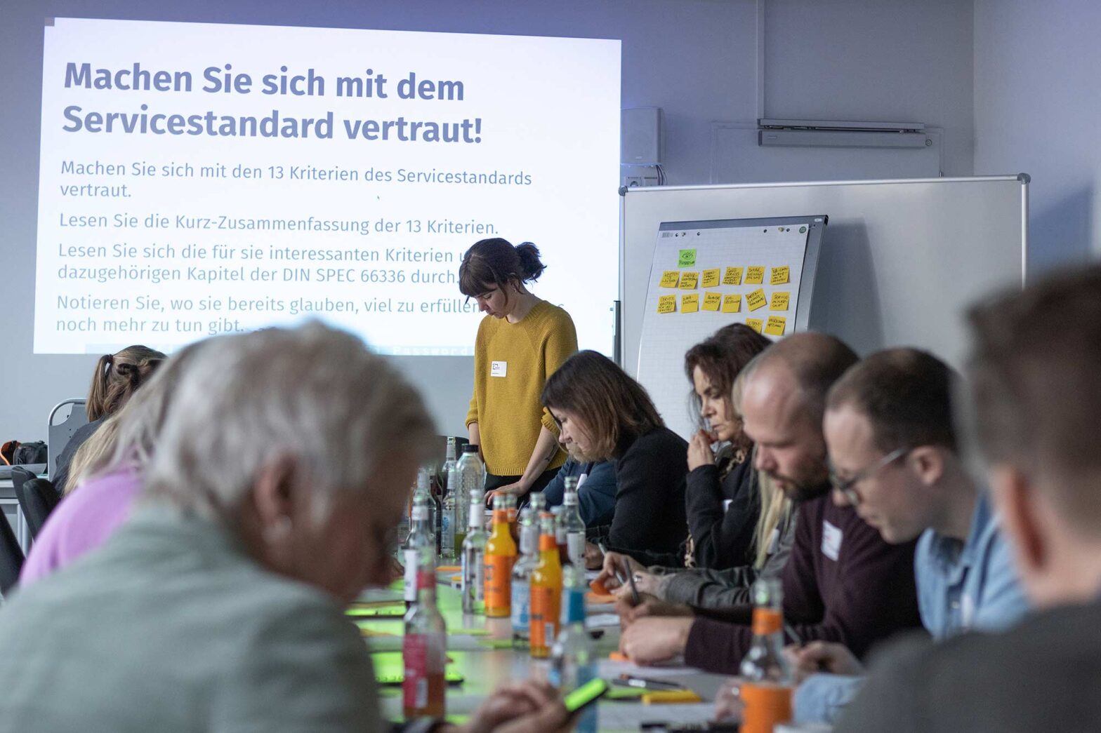 A group of people sitting around a long table in a workshop, they are focused and writing sticky notes, a large projected slide says: “Make yourself familiar with the Service Standard!”