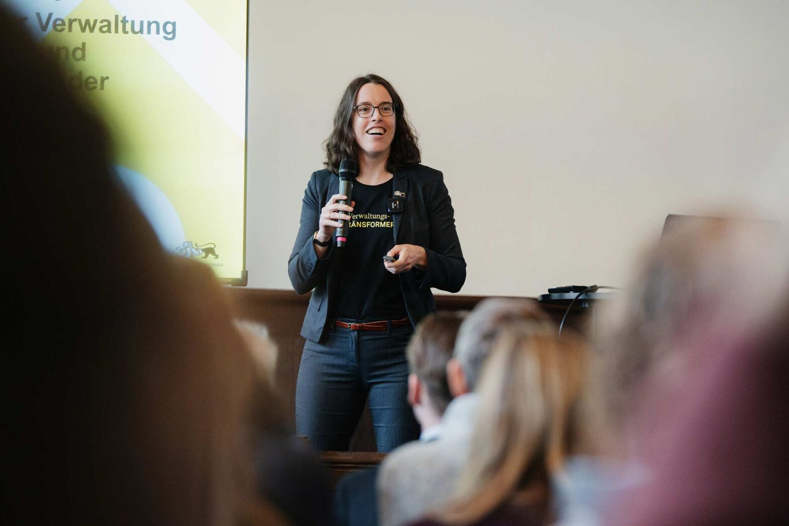 Ein Frau in Blazer über einem T-Shirt auf dem ›Verwaltungs-Tränsformer‹ steht, hält ein Mikrofon in der Hand und spricht vor einer Gruppe sitzender Menschen, sie lächelt und schaut das Publikum an