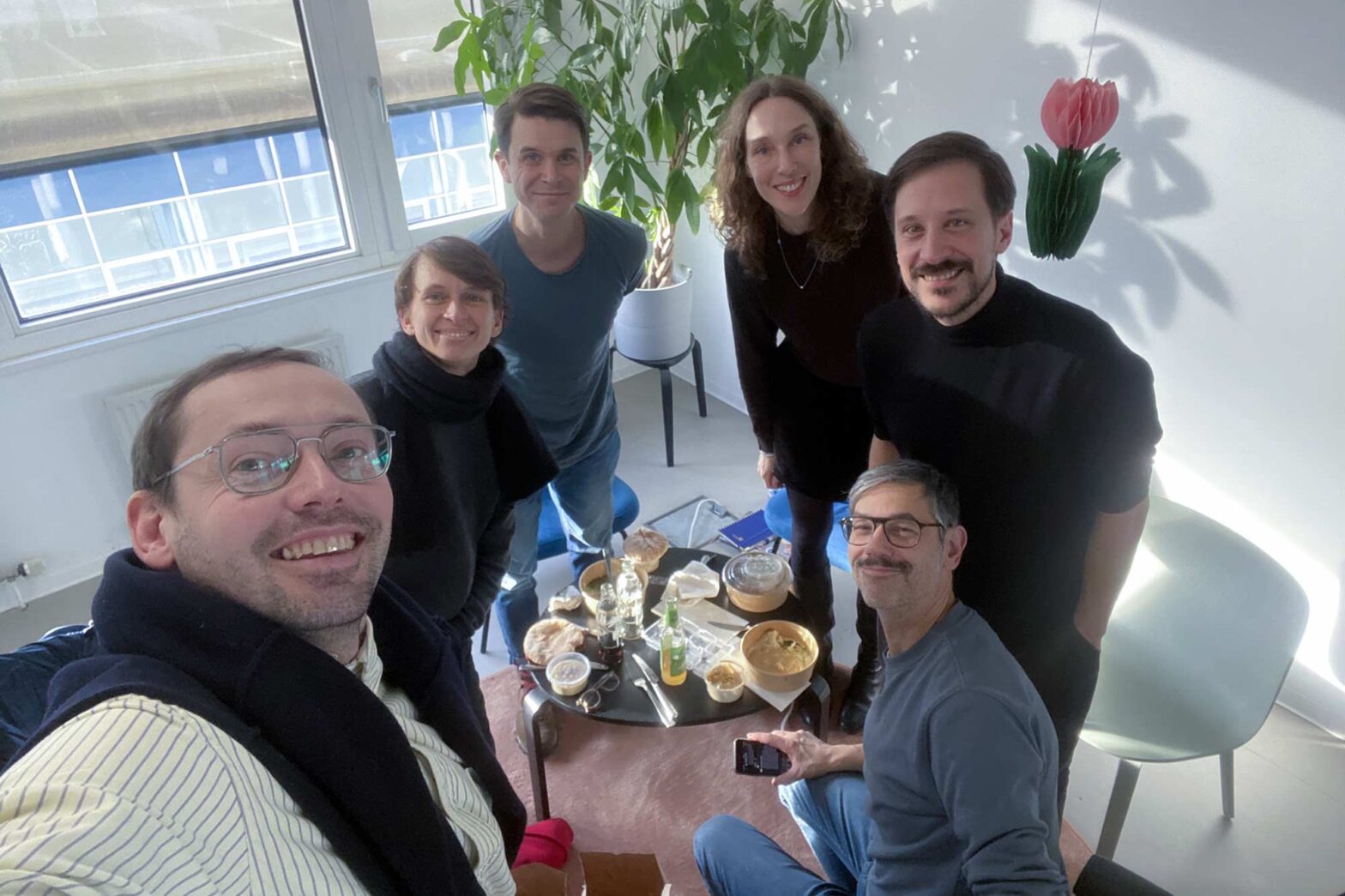 A selfie with 6 people in their 40s, all light-skinned and smiling into the camera; they stand and sit around a small table with takeaway food in a modern office setting