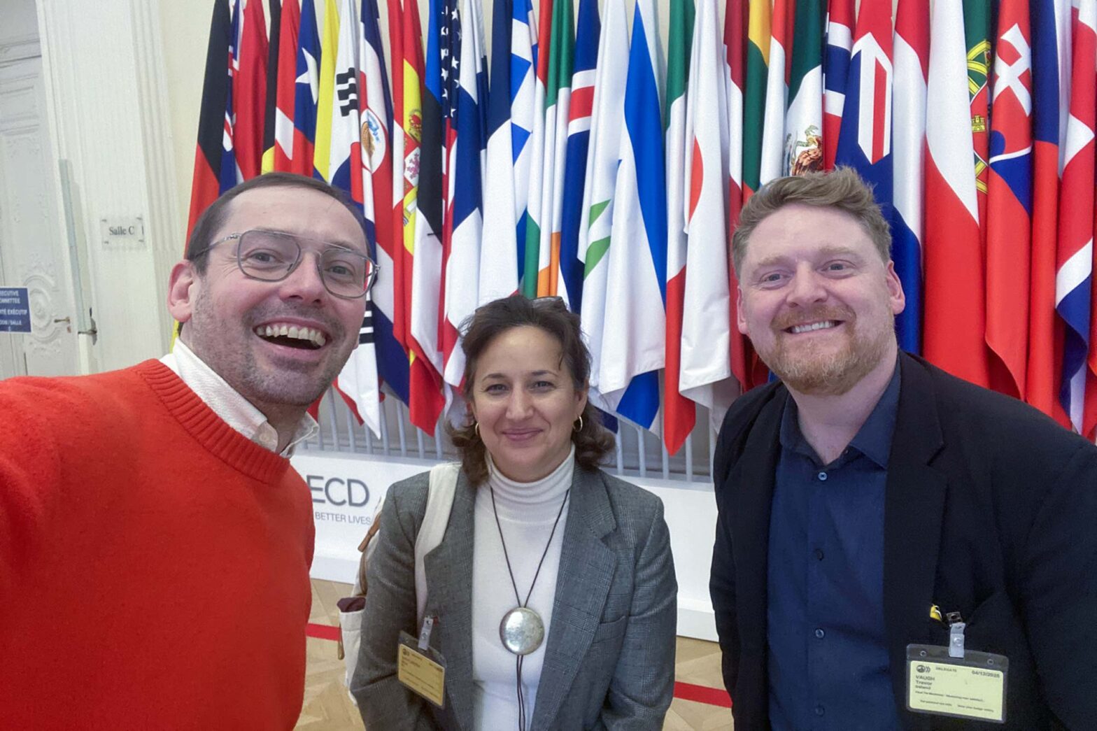 Selfie of 3 people, 2 men and a woman in a formal setting with dozens of flags of various countries hanging behind them, all 3 are smiling into the camera