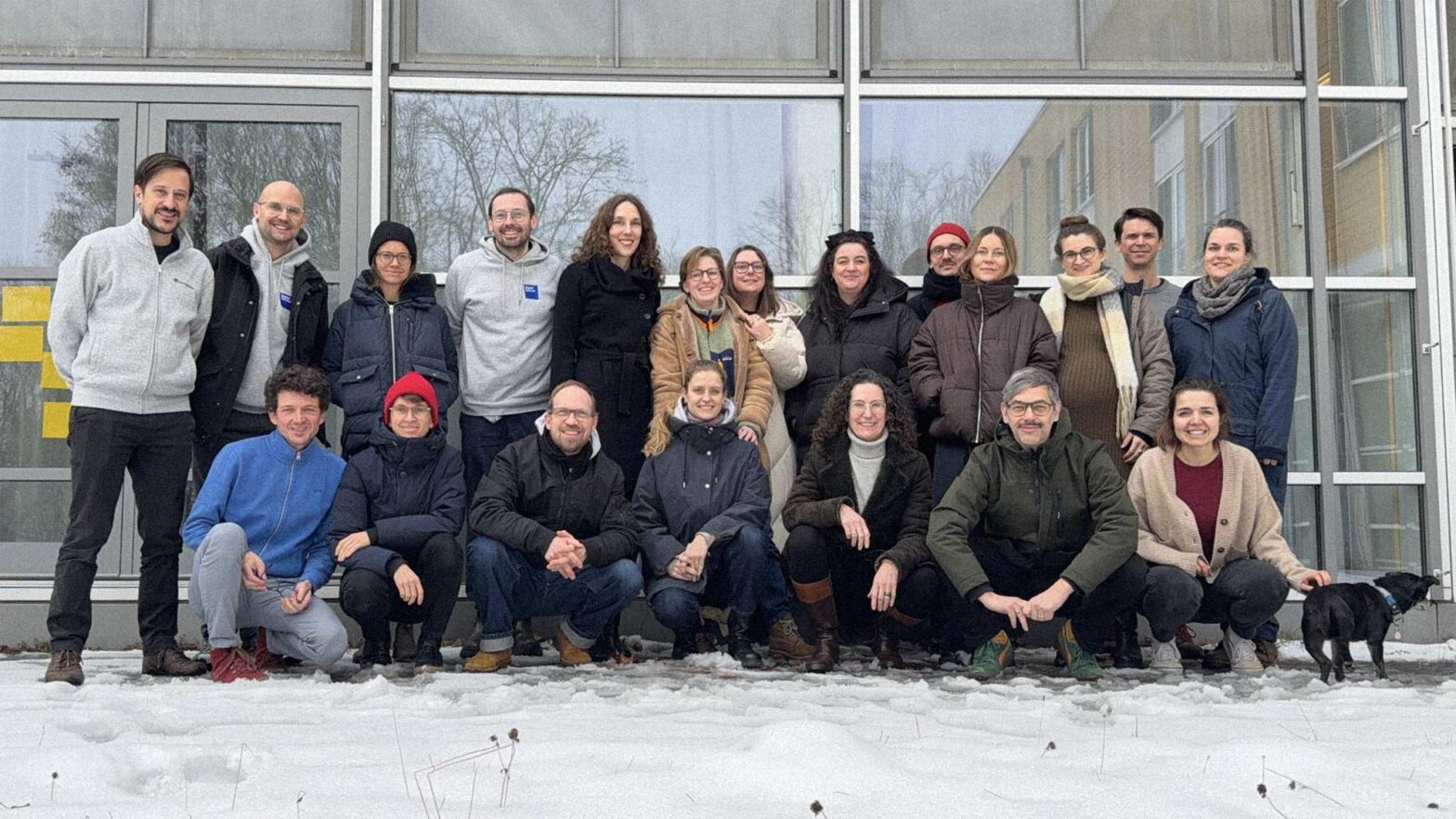 Group picture of 20 people and a small black dog standing together in 2 rows in a snowy winter setting, smiling in front of glass facade