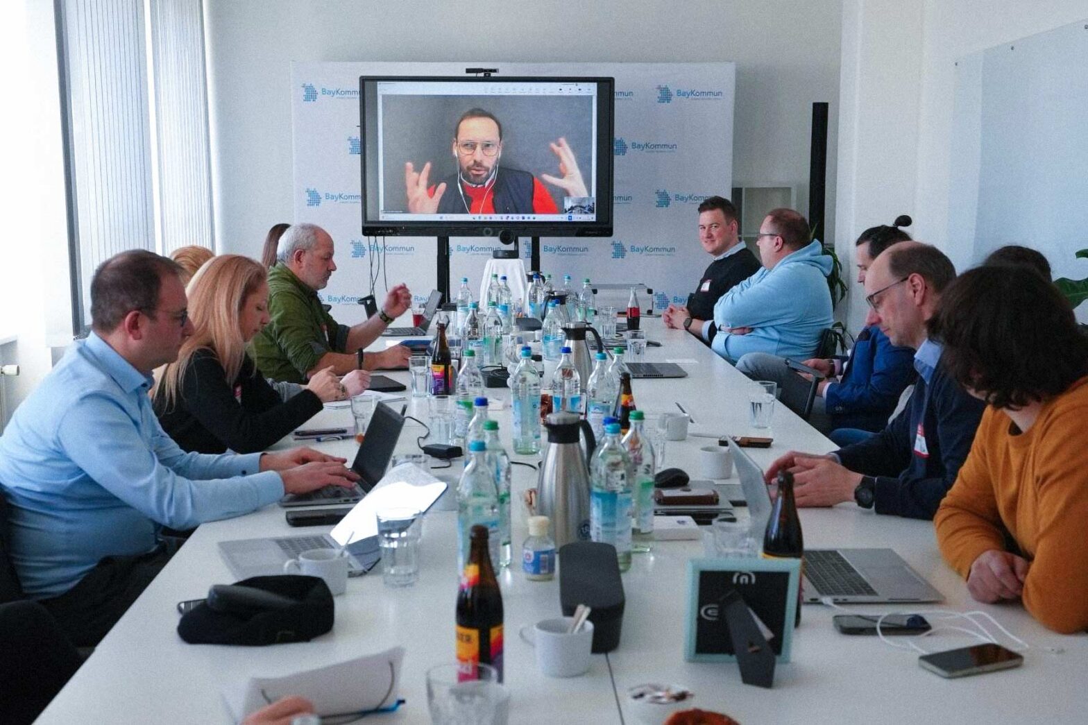 Group of about a dozen people in a meeting room sitting around a long table; a man gesticulating with hands is shown on a big television at the back of the room