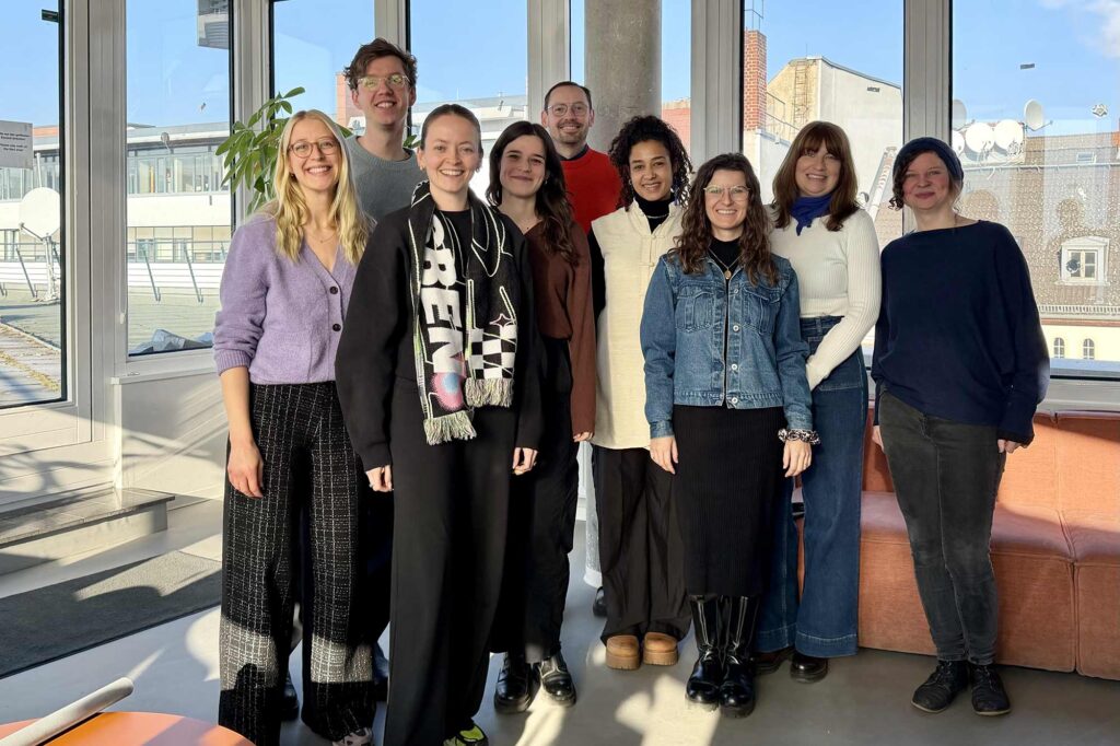 A group picture of 9 people, standing together in casual outfits in a modern office setting – 7 women and 2 men, between the late 20s and mid-40s, all are smiling