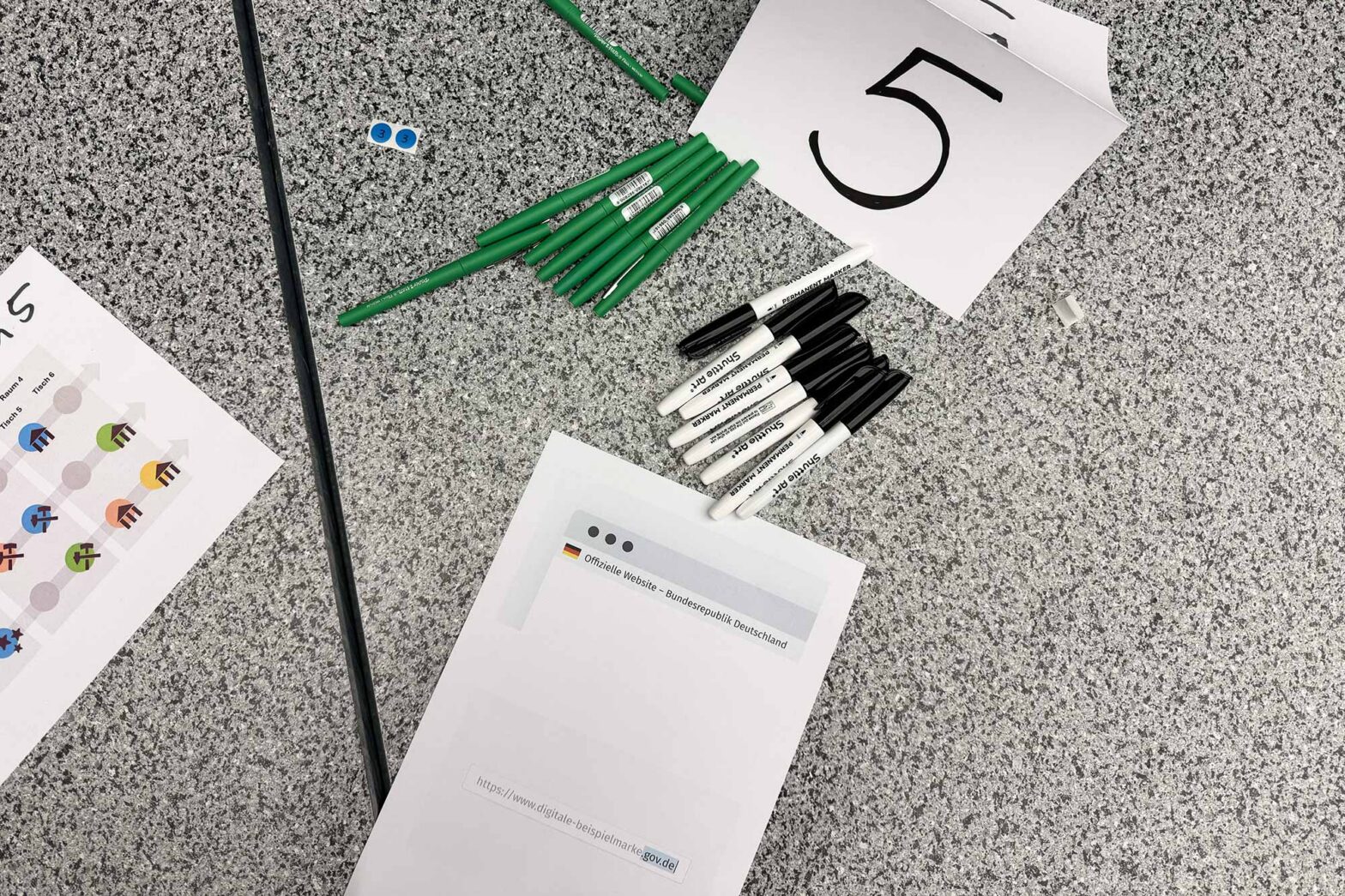 Top down view on a textured table during a workshop with 7 green pens and 8 thick markers laying next to a folded paper with a big 5 on it; 2 sheets are on the table