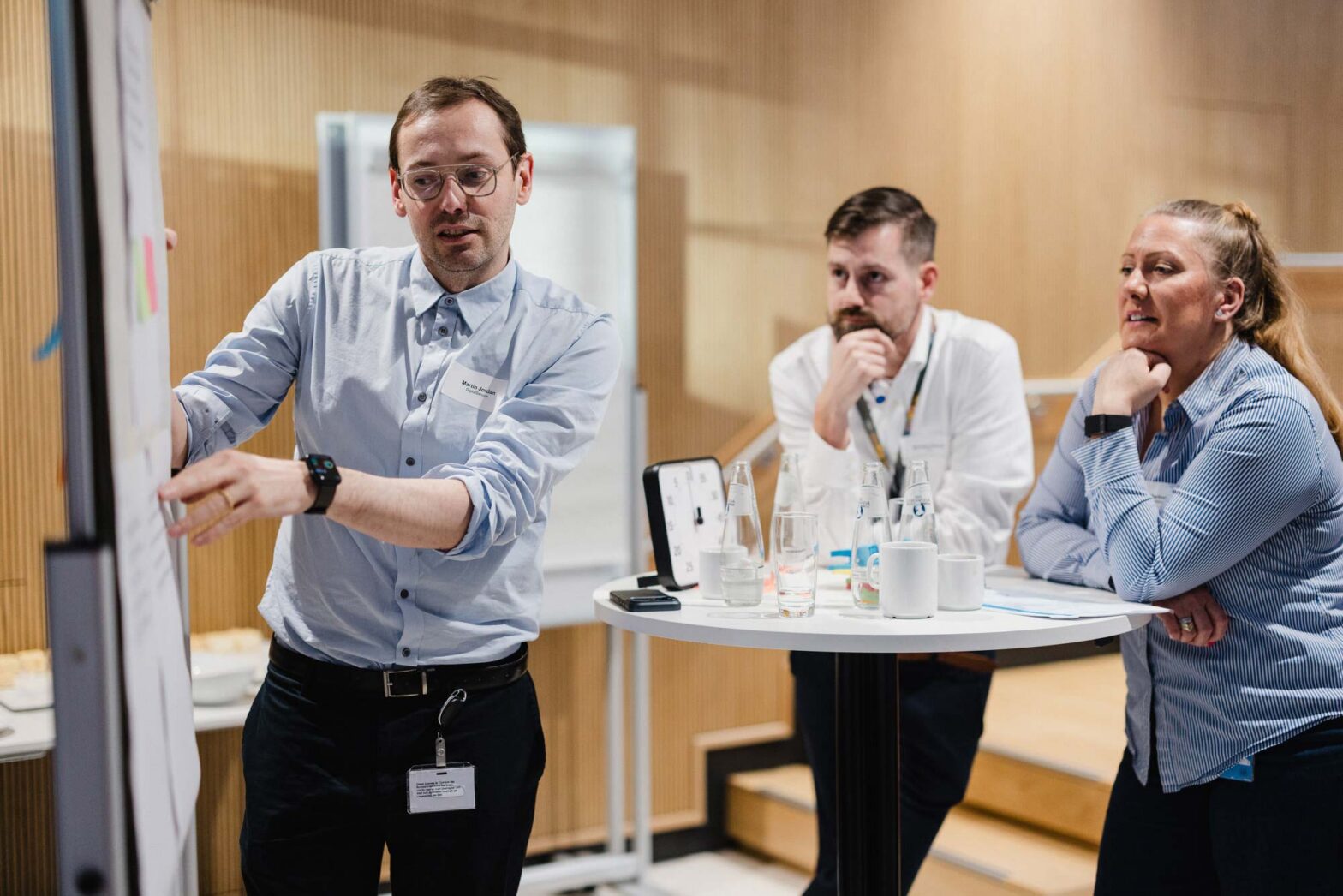 3 people in a business workshop setup: 2 men and a woman, all wearing light-colour shirts; 1 of the man is standing next to a flipchart board placing some sticky notes, the other two people looking at him with attention