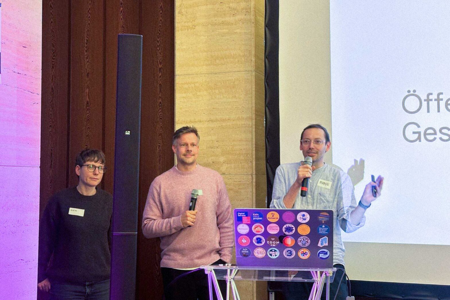 3 people presenting in a semi-casual setting, 1 woman and 2 men; one of the man holds a microphone, standing behind a lectern with a laptop computer covered with colourful stickers.