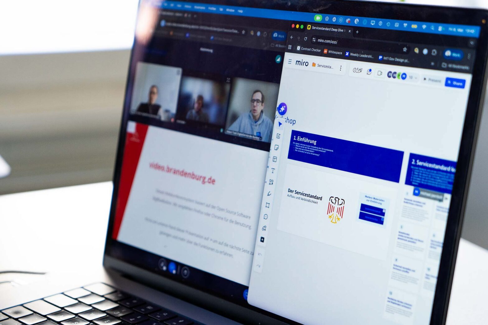 Close-up of a laptop computer screen showing two windows next to each other: 3 people in a video call with a shared slide displaying a web address on the left and a virtual whiteboard with blocks of text and graphics on the right
