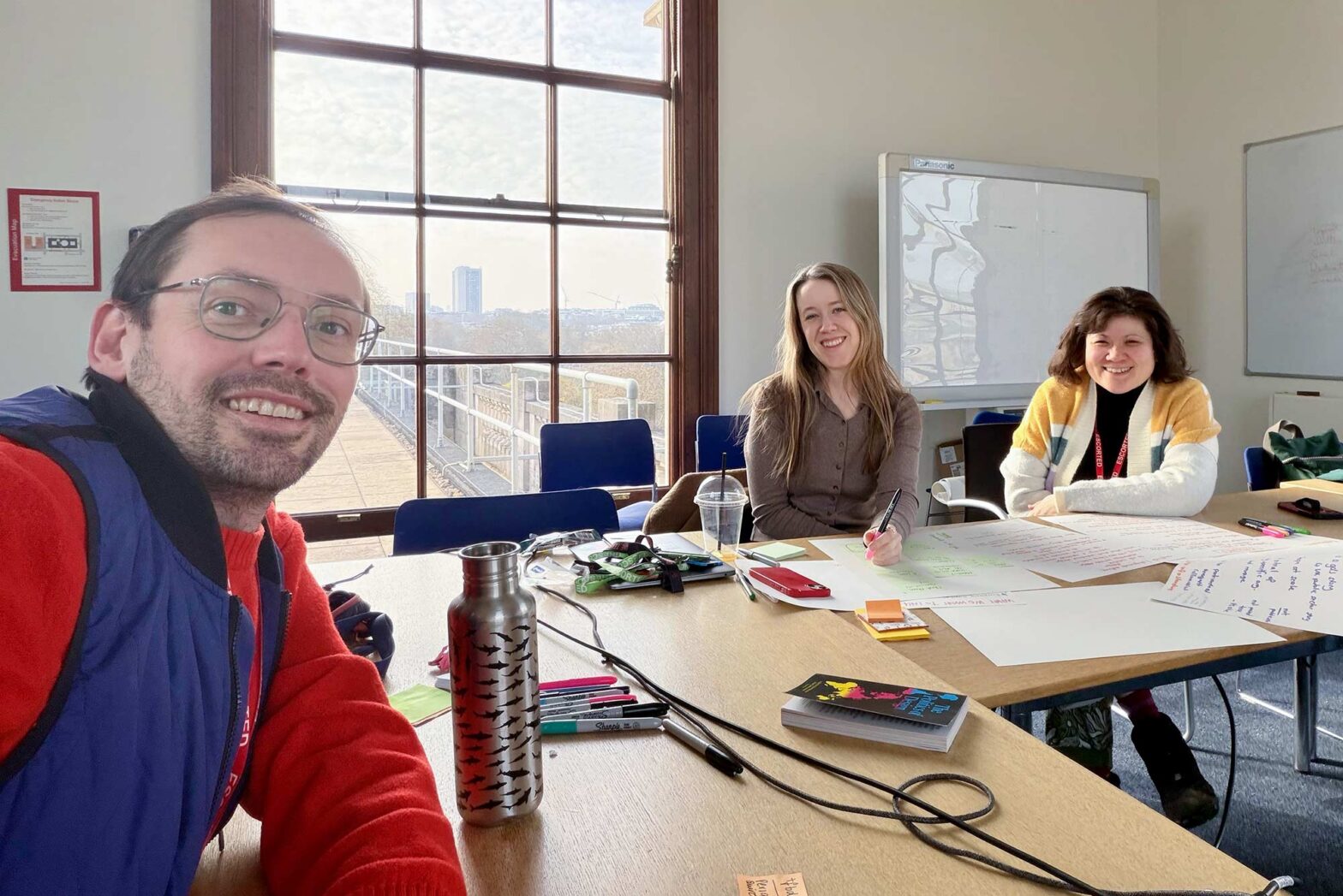 A selfie of 3 people – a man and two women – in an office setting with a big window in the background; notes, sheets and pens are on the table they sit around; all 3 are smiling into the camera