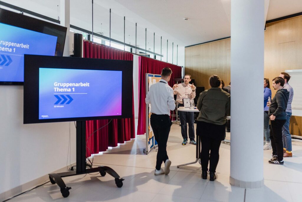 A modern government building with wooden panels, white walls, a column – in the foreground 2 TV sets displaying the same slide saying ‘Group work, topic 1’; in the background is a group of 7 people in a workshop setting with a large pin broad, a timer and high table with colourful sticky notes; one man is talking using his hands, the others are looking at him; 
