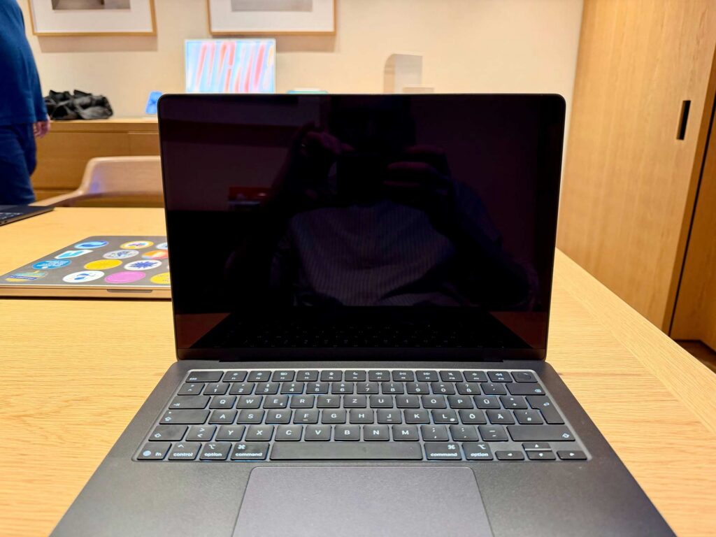 A dark silver laptop computer with a black screen is standing on a wooden table with another laptop with colour stickers behind it; on the wall are some wooden frames with a wooden sideboard below