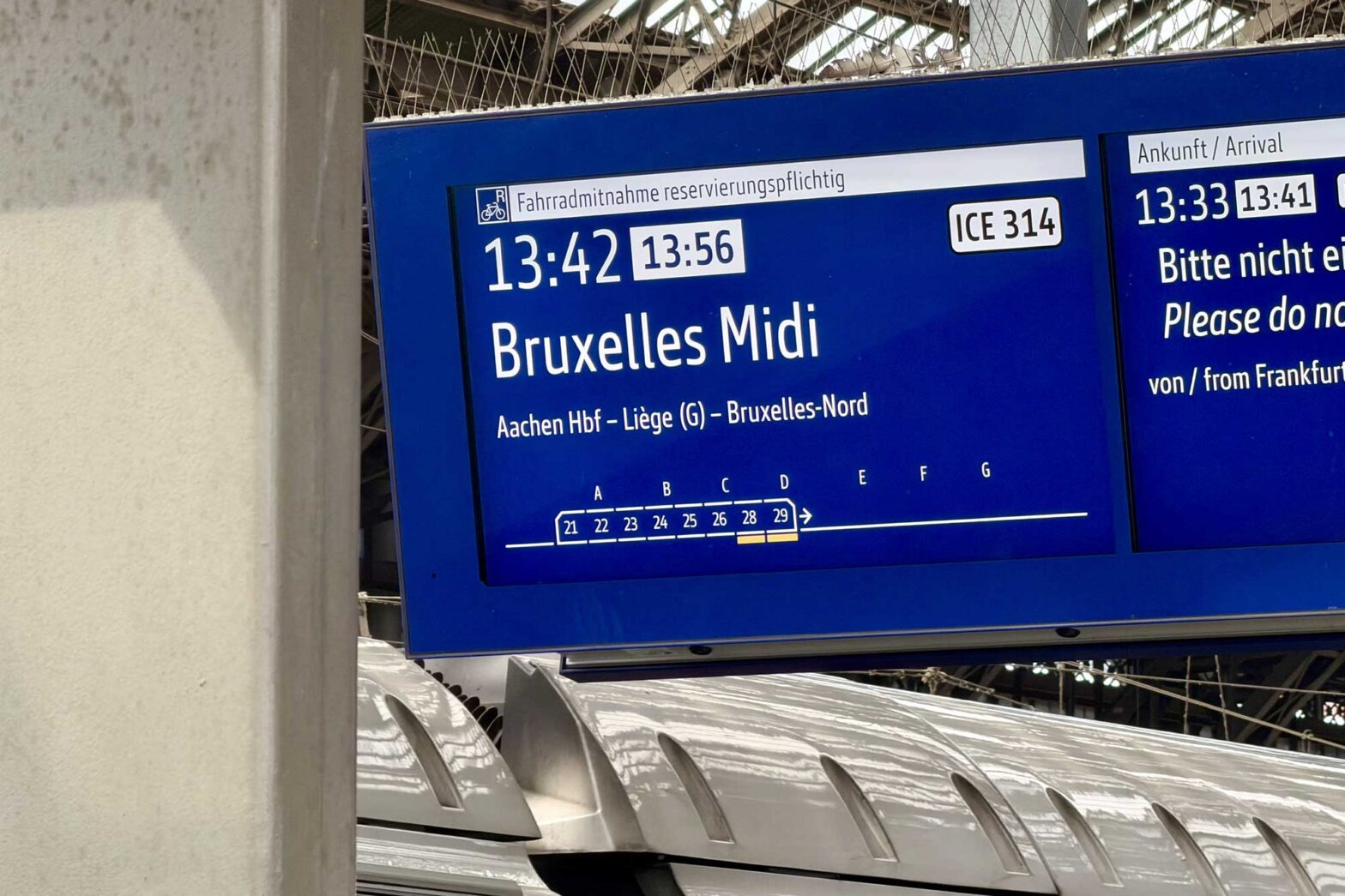 A sign in a German rail station displaying a 13:42 connection to Brussels Midi station, delayed by 14 minutes, behind it, the roof of a German speed train is visible