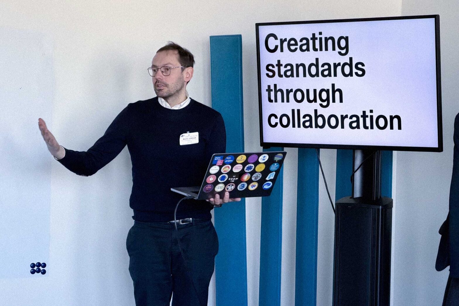 A light-skinned middle-aged man in a casual business outfit, talking while holding a laptop computer covered in colourful stickers next to a TV set, displaying a big headline: Creating standards through collaboration