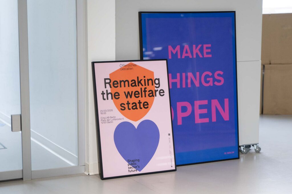 2 posters in frames leaning against a wall and cupboard; 1 poster shows a big purple heart and orange shield, saying ‘Remaking the welfare state’; the other one says: ‘Make things open’