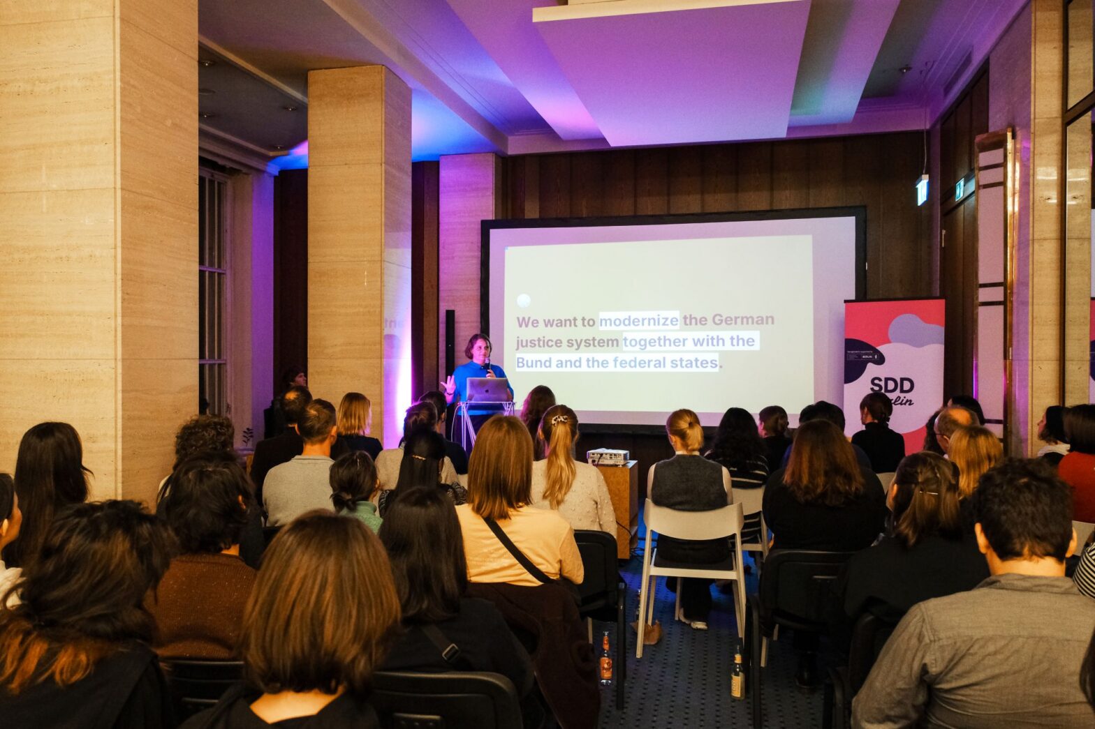 An event space with a woman presenting in front of a larger group of seated people, behind her is large projected screen, saying ‘We want to modernize the German justice system together with the Bund and the federal states.’