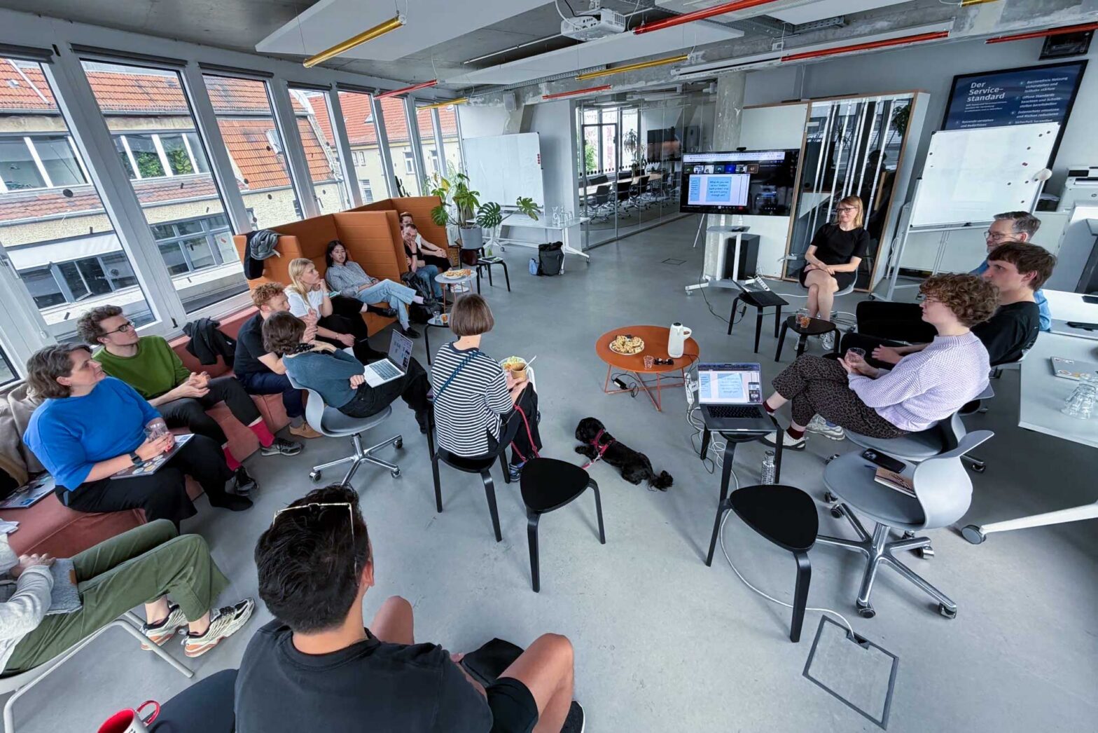 A group of about 20 people sitting in an open office space on sofas, chairs, and stools with a TV set standing in the background