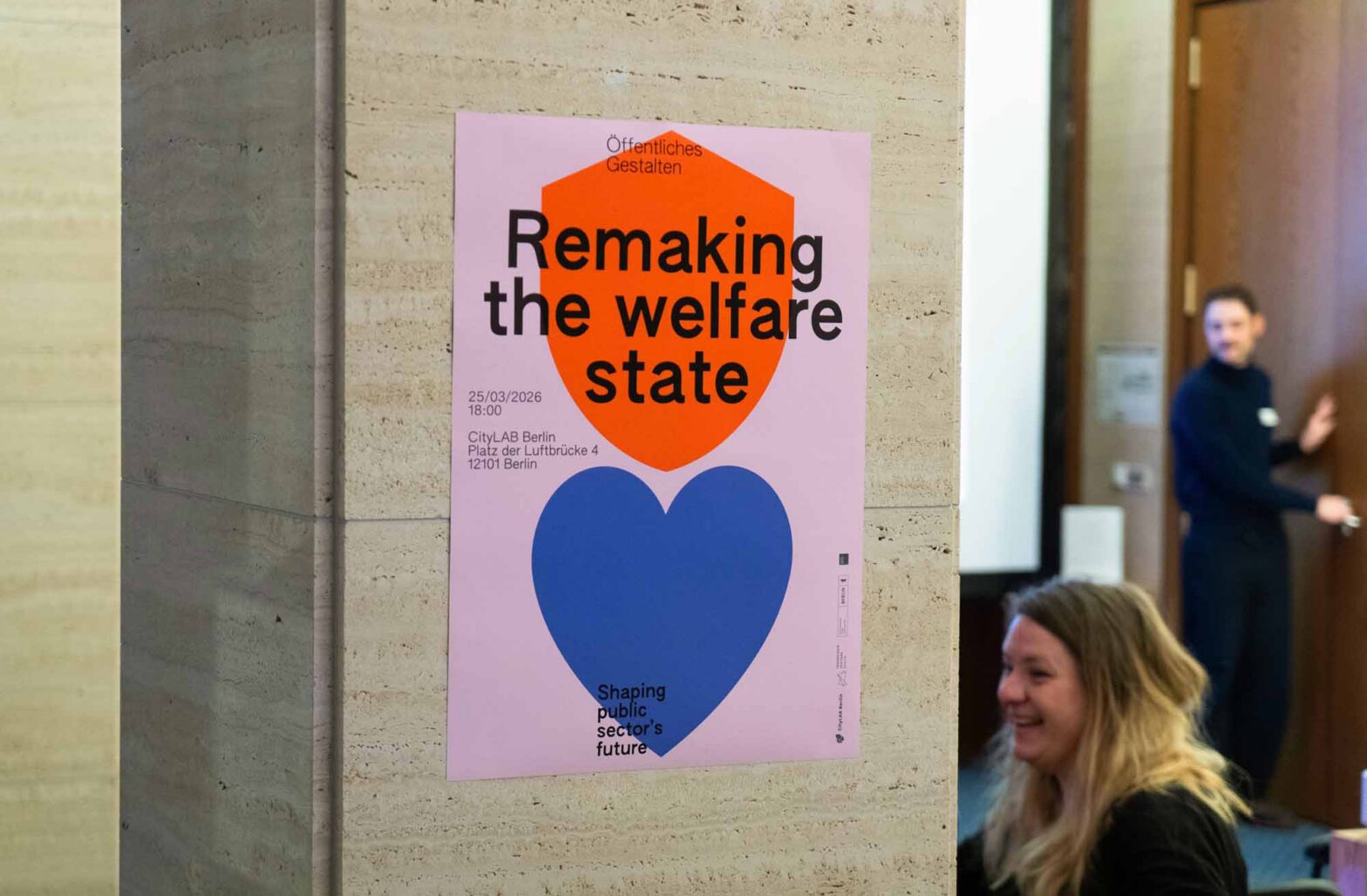 A poster attached to a marble column showing a large purple heart and an orange shield, with the words ‘Remaking the welfare state’; next to it are women smiling, sitting, and, further behind, a man at a wooden door.
