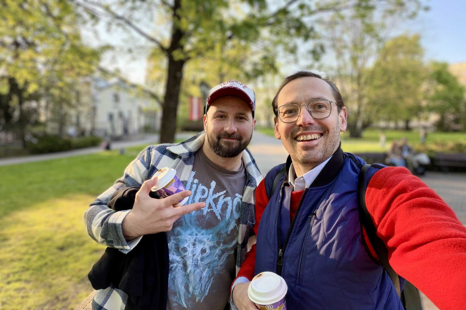 Activate to view larger image, Selfie of 2 light-skinned middle-aged men in a park setting, both smiling, holding a cup of takeaway coffee