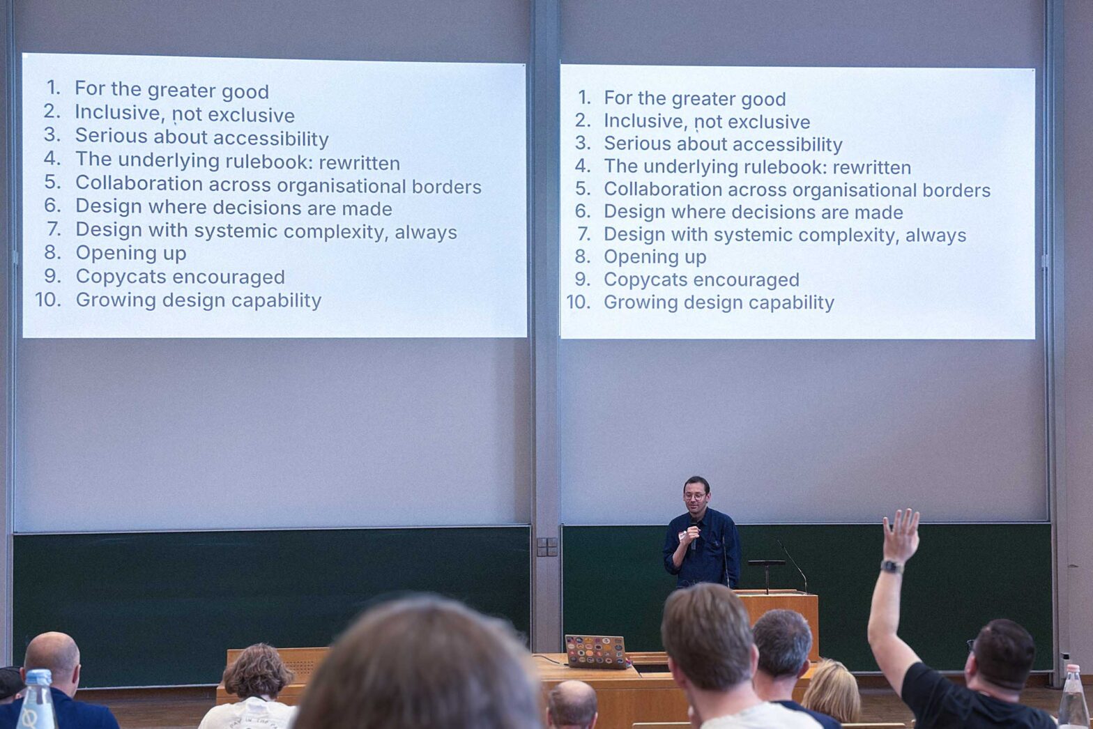 A middle-aged man in a dark shirt presenting in a seminar hall in front of seated people, one of them is raising their hand; behind the presenter, standing at a lectern are 2 identical projected slides with a list of 10 points on design in government