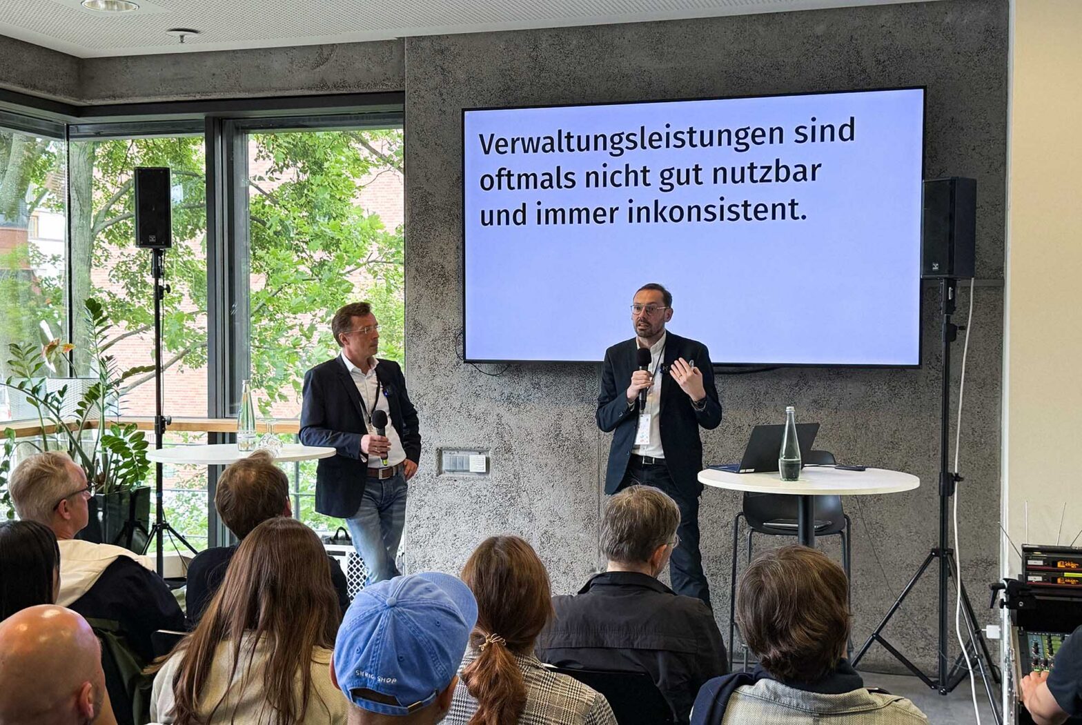 2 mittelalte Männer mit heller Haut in Sakkos und weißem Hemd sprechen vor einer Gruppe sitzender Menschen in einem Tagungsraum; hinter ihnen ist ein großer Bildschirm, auf dem zu lesen ist: ›Verwaltungsleistungen sind oftmals nicht gut nutzbar und immer inkonsistent.‹