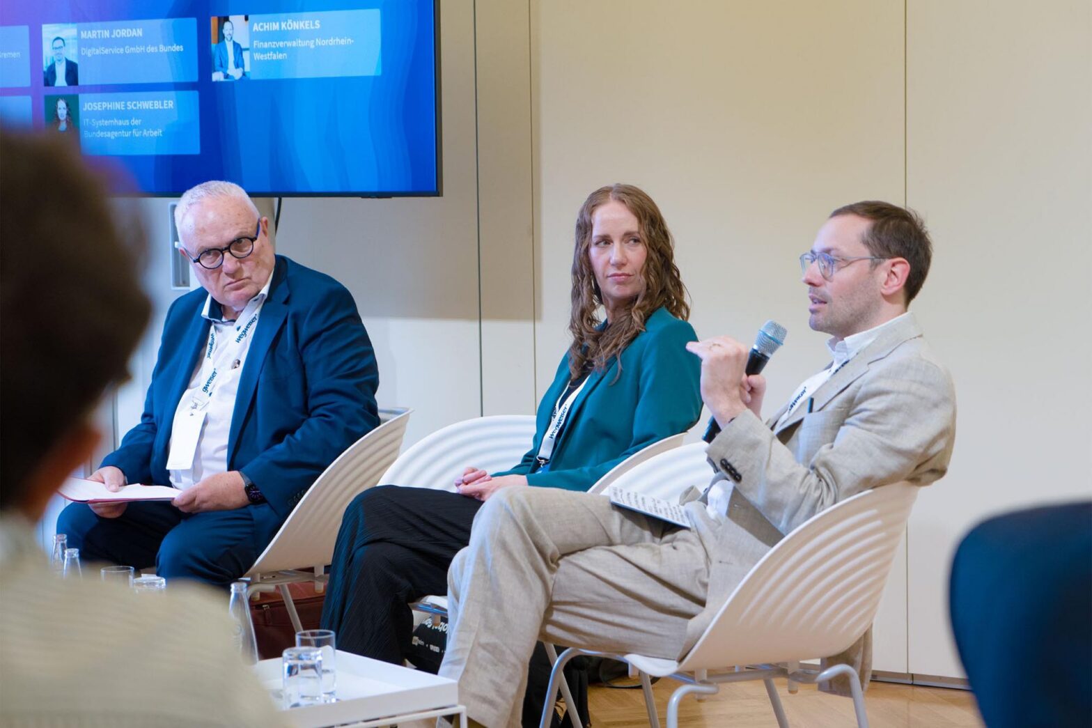 3 seatedpeople during a panel discussion – an older man with grey hair, a middle-aged woman and a middle-aged man; all have light skin and wear formal clothes, the younger man wears glasses and holds a microphone; people are sitting in front of the panellists