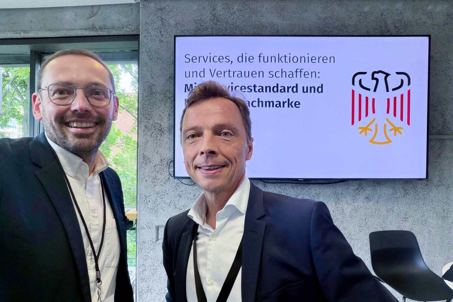 Selfie of 2 middle-aged light-skinned men standing in front of a large TV set with a big eagle symbol of the German government