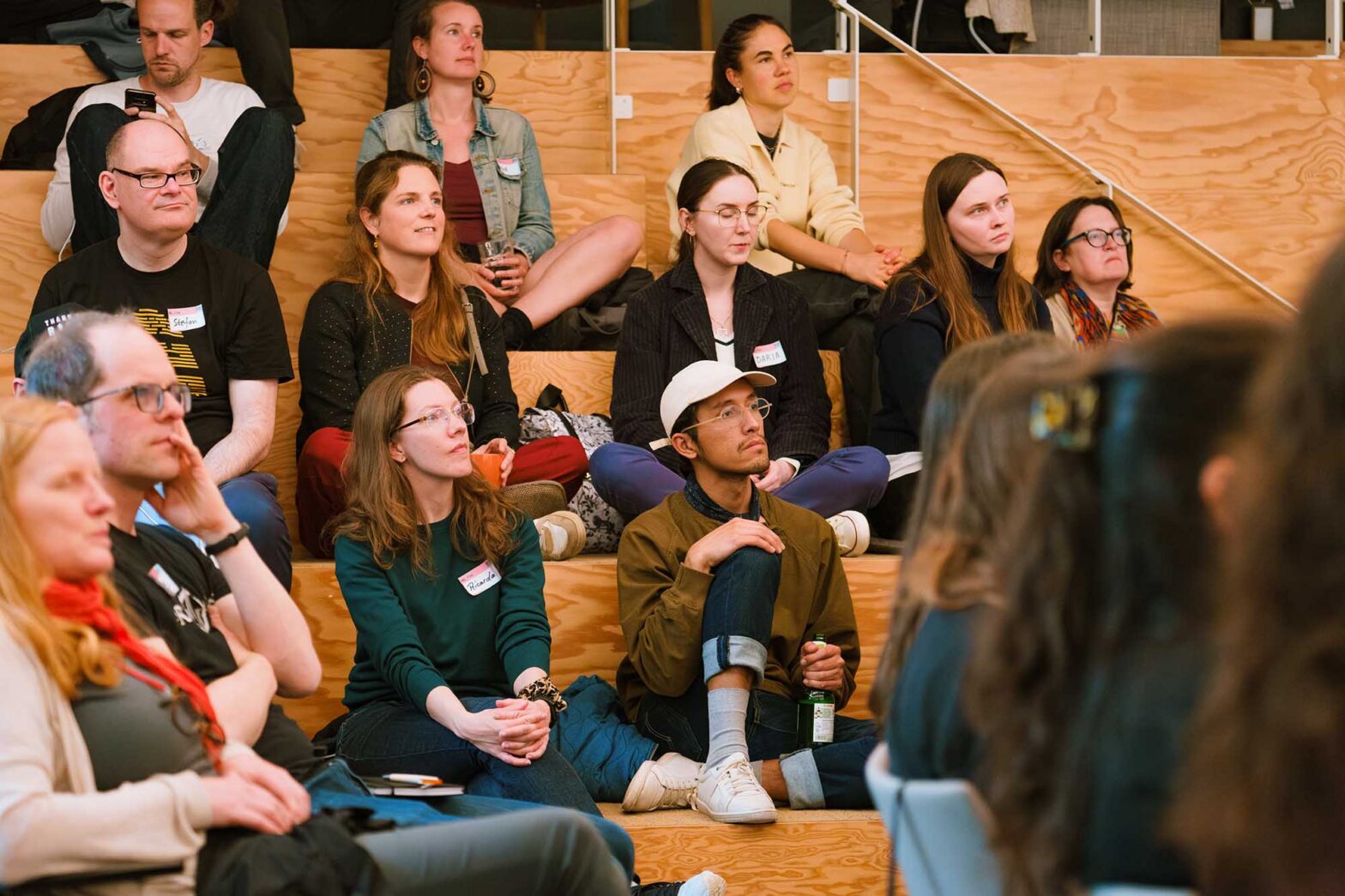 Over a dozen people sitting in a community space with wooden stairs, listening and looking in the same direction, they are wearing casual outfits