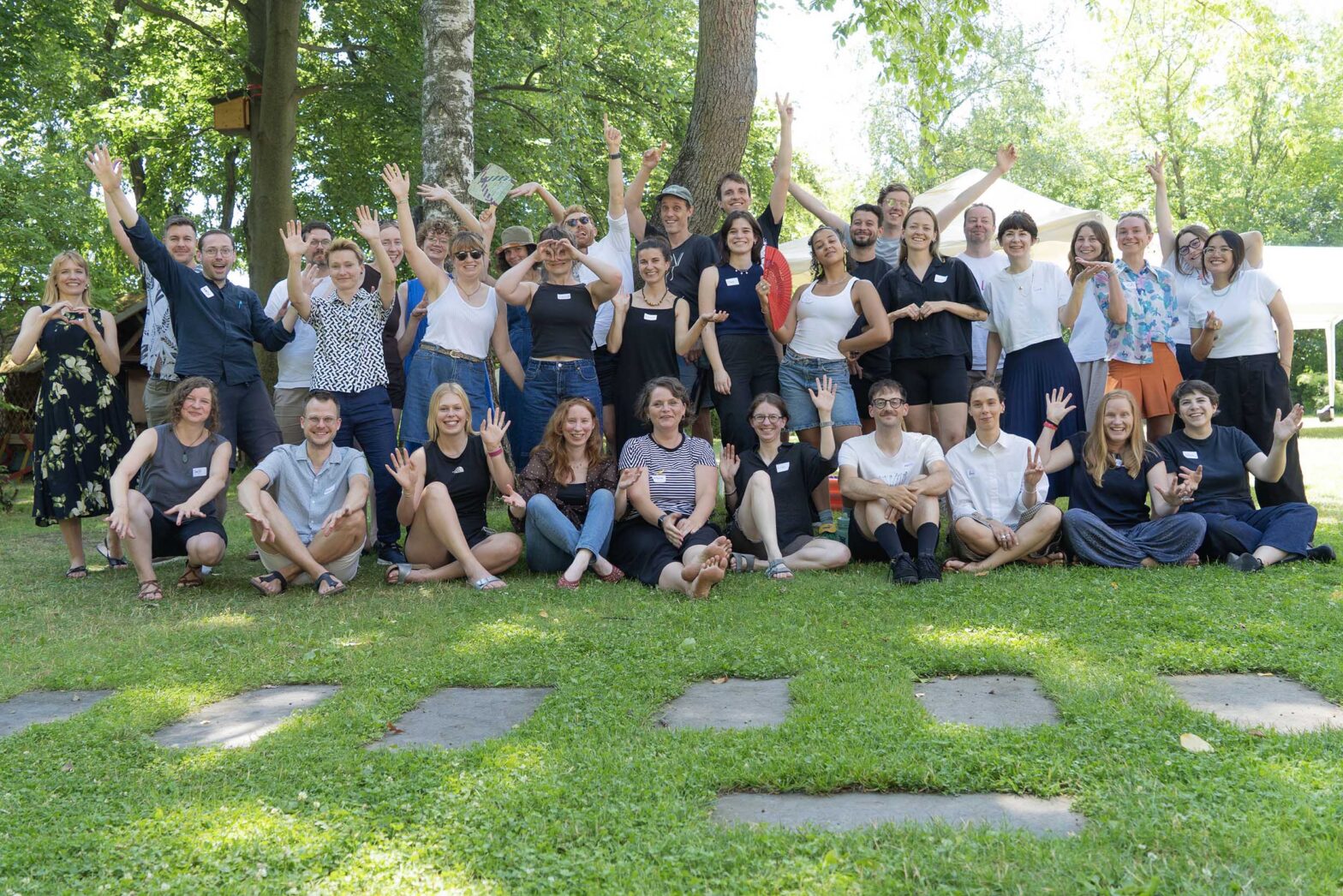 A group of 35 people posing for a picture in an outdoor garden or forest space, people are cheerful, raising their hands, smiling, all wearing casual summer outfits
