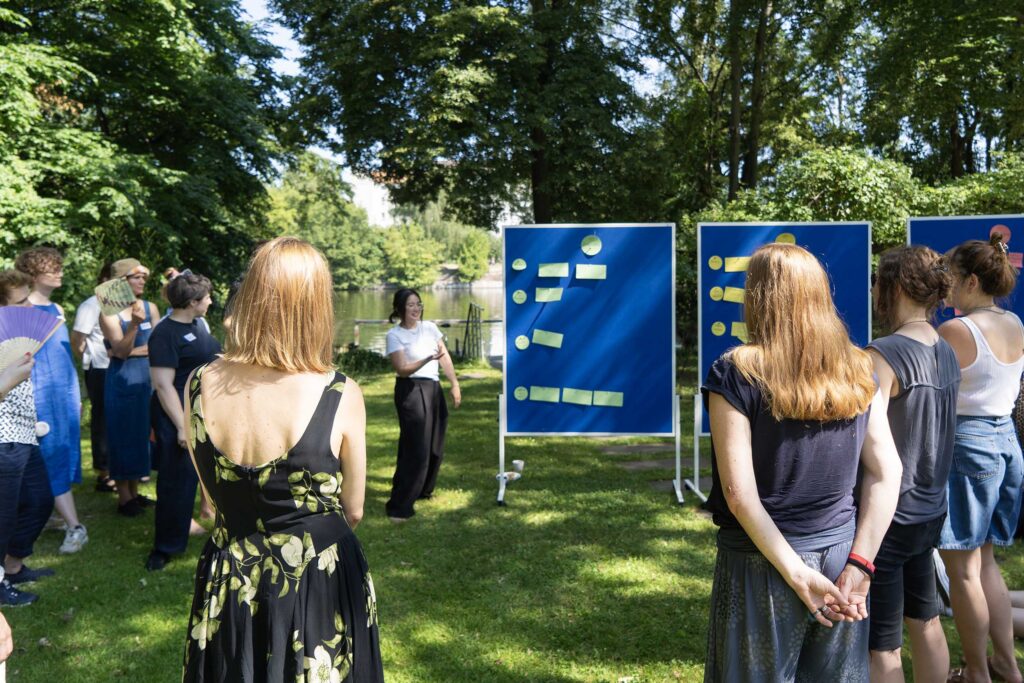 A group of about a dozen people is standing in an outdoor space with office pinboards with pinned cards in various colours, people wearing summer clothes and holding fans, one woman of asian decent is speaking; she is smiling, the others are looking towards her