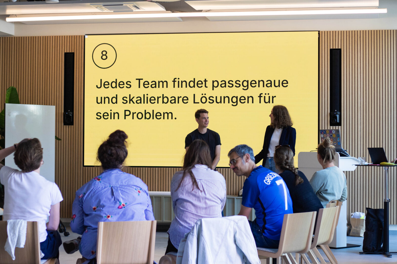 2 people, a man and a woman, presenting in front of a group of seated people in a modern meeting space with a wood-panlled wall and a large screen, which says in German: “6 – Each team finds suitable and scalable solutions for their problems”