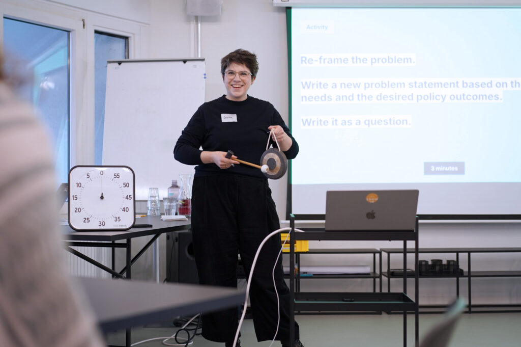 A woman with lighter skin, short hair, smiling, holding a gong, standing in a modern office space in a workshop setup; a big timer is next to her and a laptop computer; a person is sitting in the foreground, in the background is a projected slide reading: “Re-frame the problem: Write a new problem statement based on the needs and the desired policy outcomes. Write it as a question.”