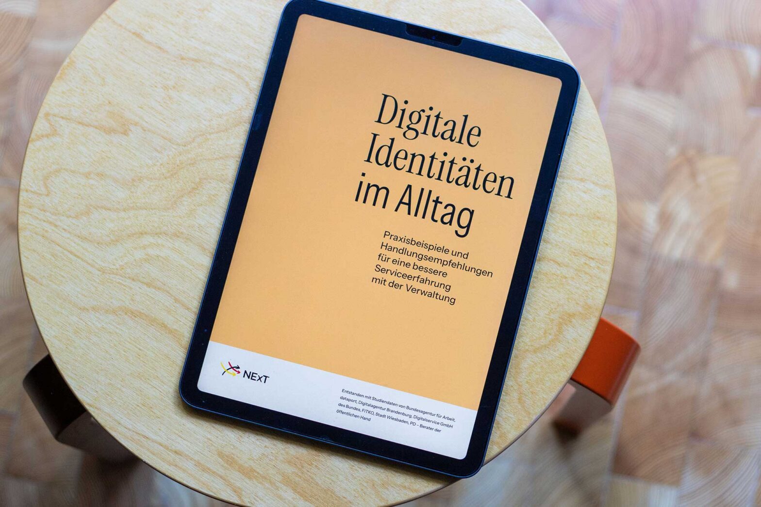 A tablet computer on a stool, displaying a cover image that reads: “Digital identities in everyday life: practical examples and recommendations for a better service experience with public administration”, with the NExT logo on it