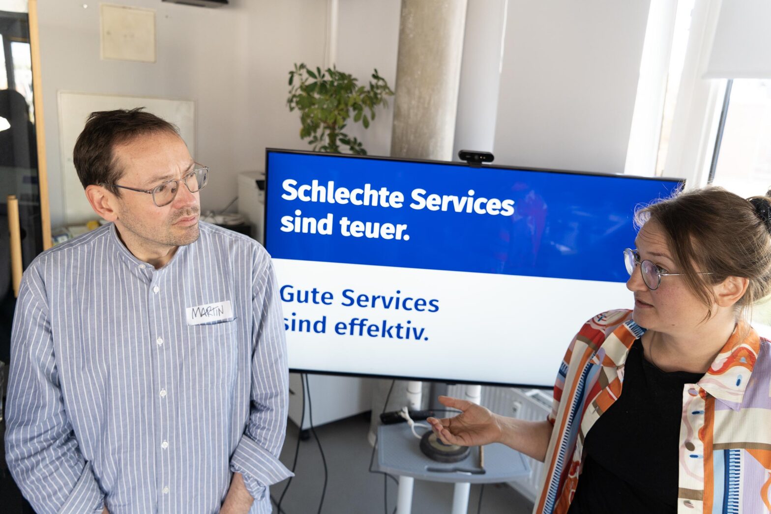 2 people – a man and a woman – talking in front of a TV set that shows a slit-coloured slide saying “Bad services are expensive. Good services are effective.” – in German; the woman seems to be talking