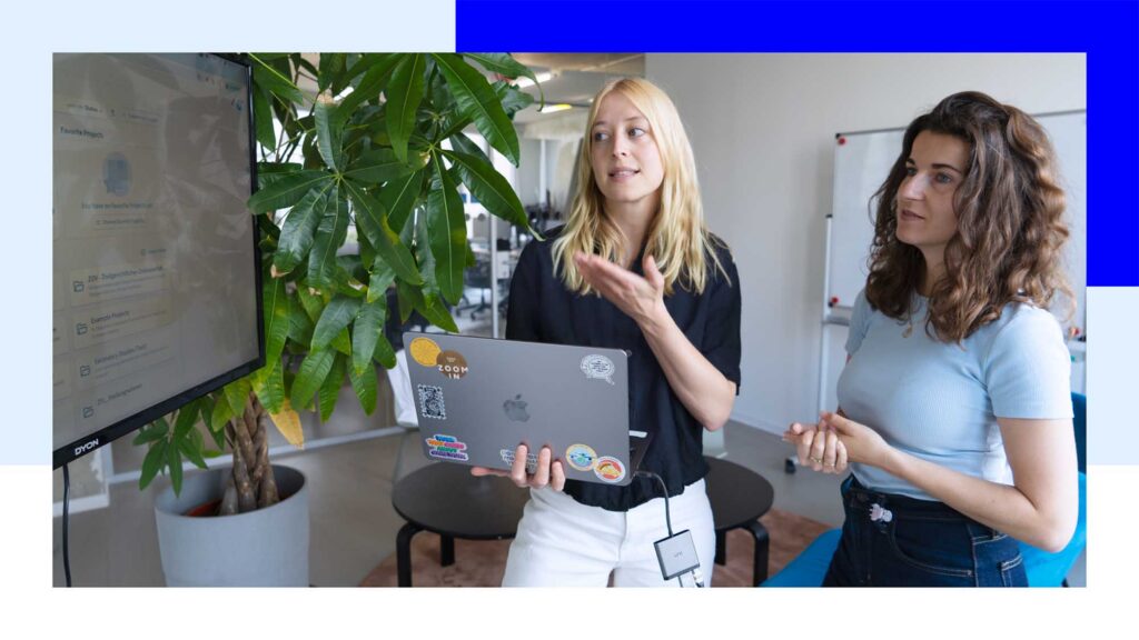 Two young-ish women with lighter skin in semi-casual outfits both look at a larger TV screen displaying a simple software interface; one of the women holds a laptop computer with various colourful stickers; the women stand in a modern office environment next to a large green plant and in front of a white board