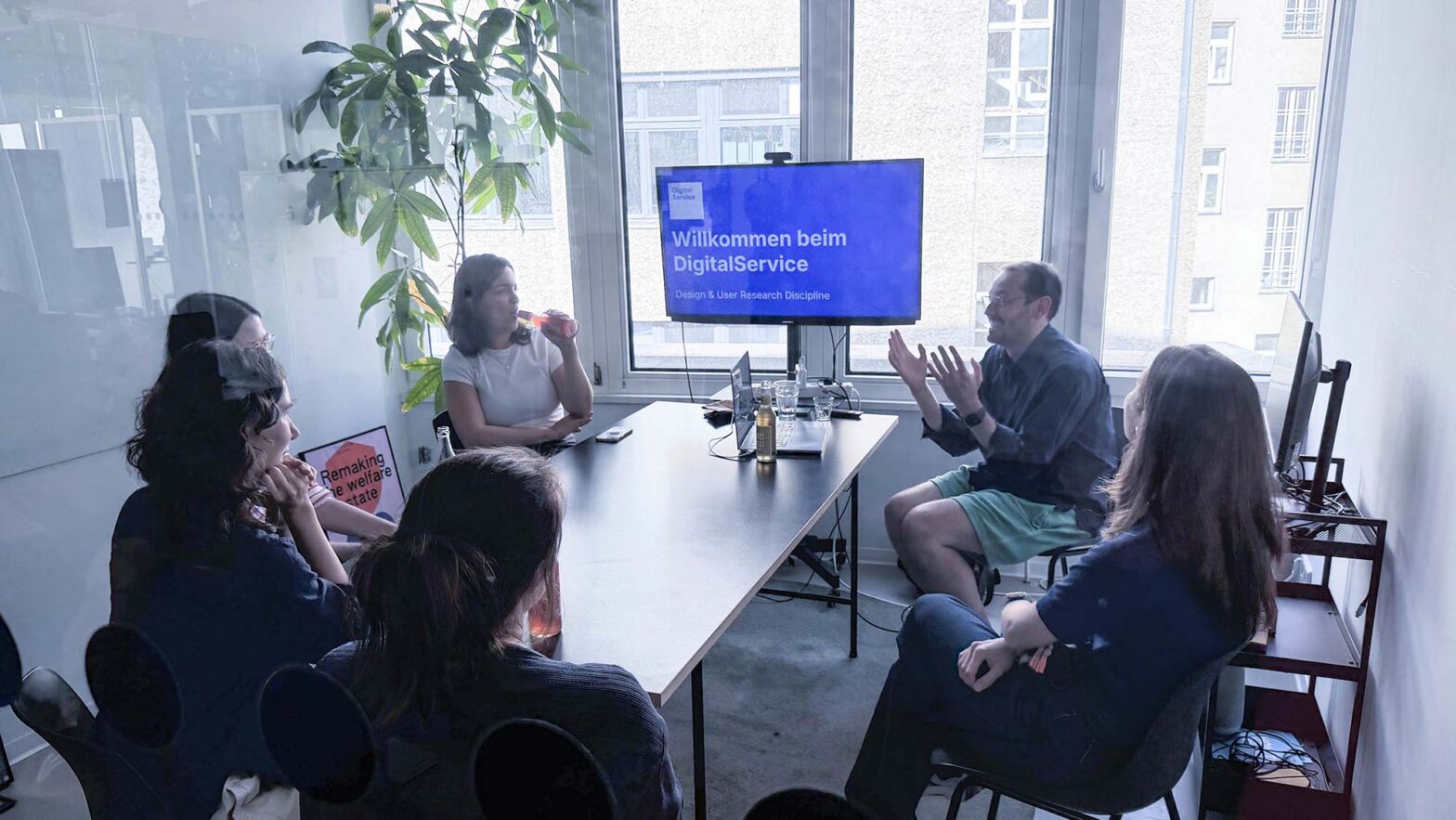 A man and 5 women in a small modern glass-window meeting room with plants in the background and a TV set at the end of the tabled they are seated around; it shows a blue screen with white text saying: “Welcome to Digital Service”
